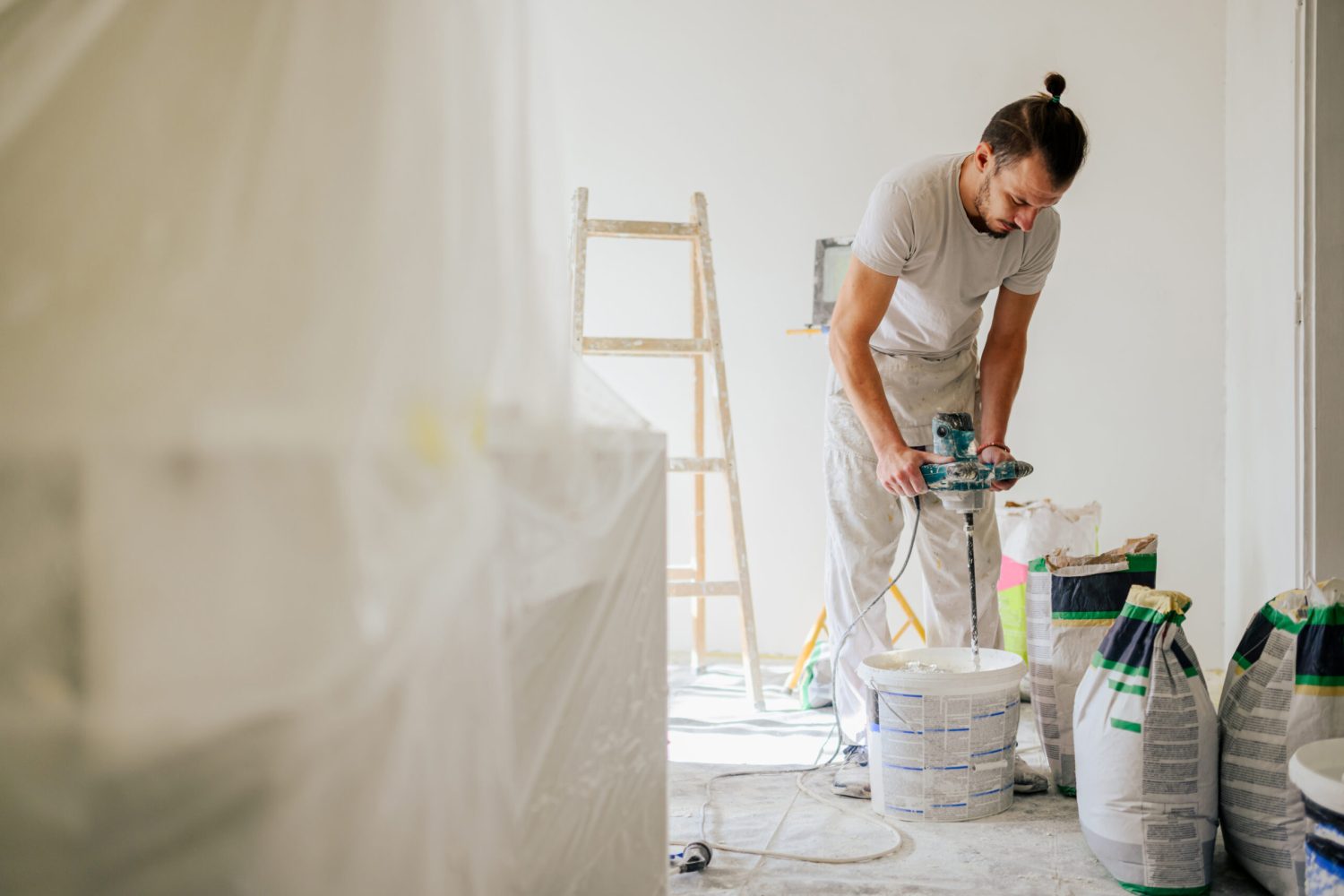 A contractor is mixing plaster with manual cement mixer while standing in a house in renovation process. A constructor is mixing and preparing plaster for skim coating and plastering walls in a house.