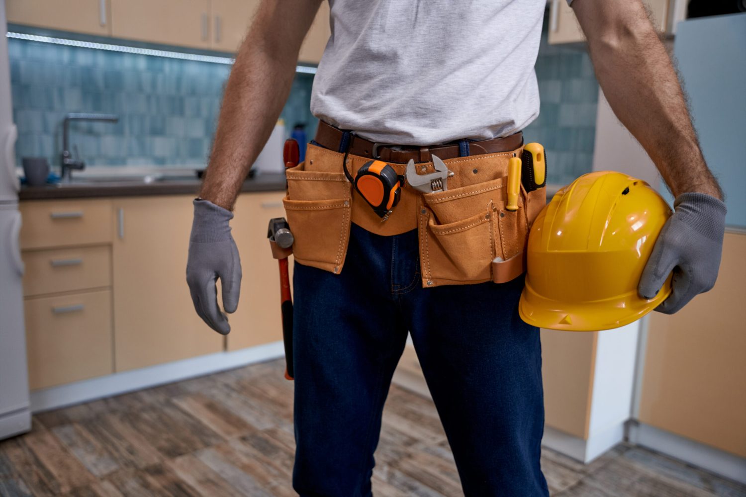 Young repairman with tools belt and hard helmet standing in kitchen, cropped. Home master concept, construction and repair